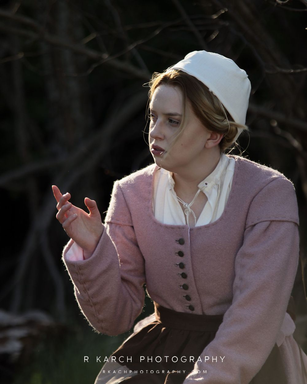 A dramatic portrait in period costume features someone gesturing while wearing a pink bodice and white cap.