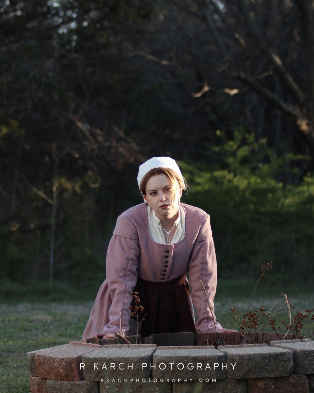 A period-dressed figure leans on a stone surface in a dusky outdoor setting.