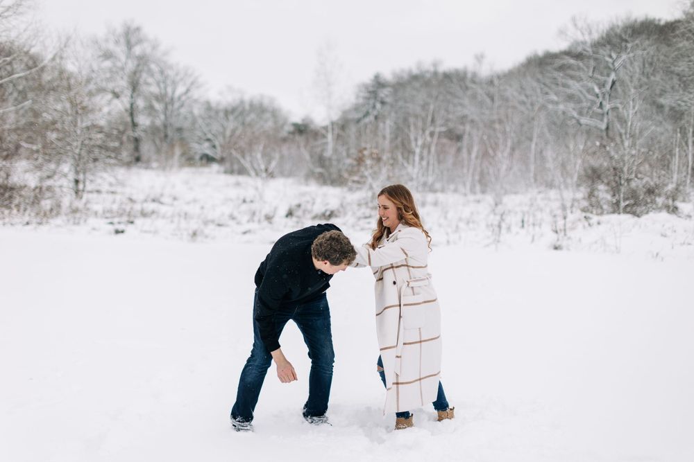 Two people play and laugh together in a snowy winter field surrounded by bare trees.