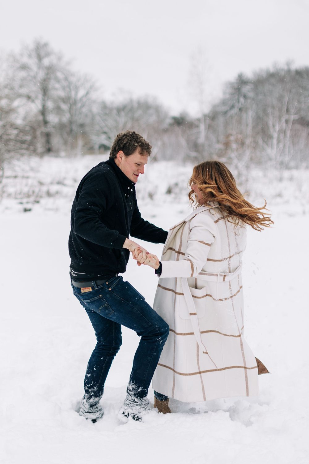 A playful couple dances together in the snow with one person wearing a cream-colored winter coat.