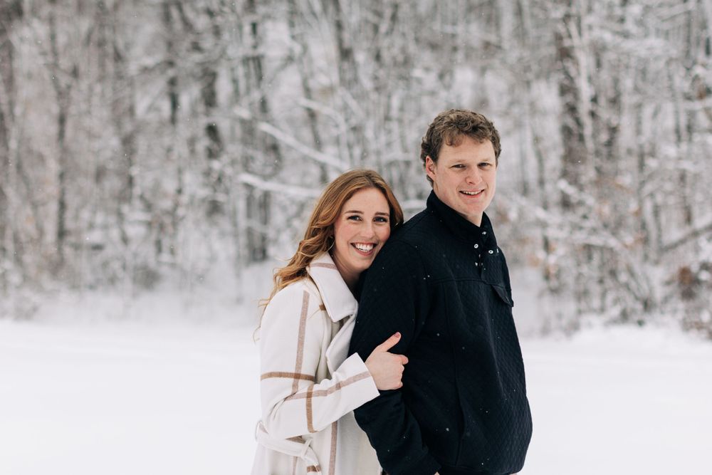 Winter engagement photo of couple in black and white coats against snowy forest background.