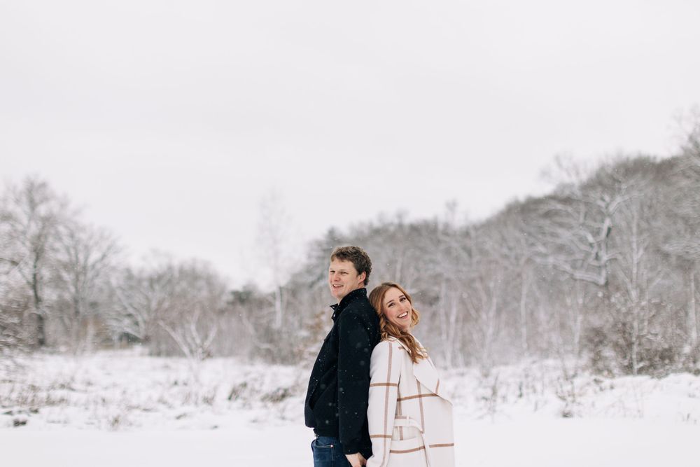A couple stands back to back in a snowy winter landscape with bare trees in the background.