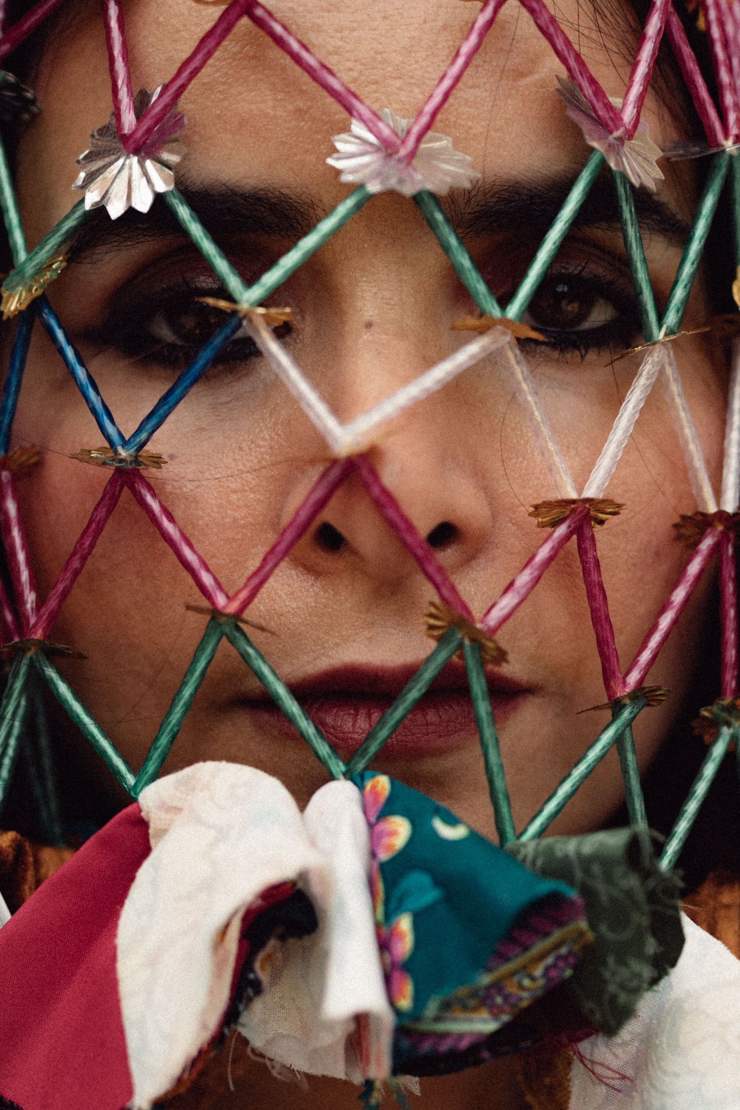 Close-up artistic portrait through colorful criss-crossed strings with small white daisy decorations and fabric elements.