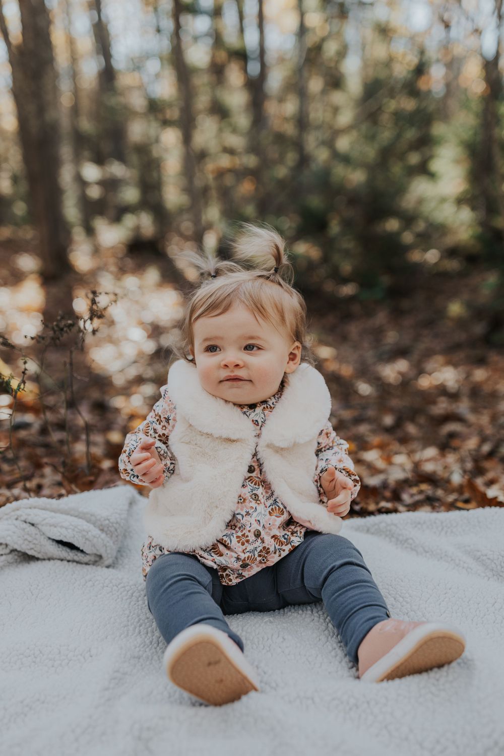 Baby wearing a white furry vest sits on blanket in autumn forest setting with warm lighting.