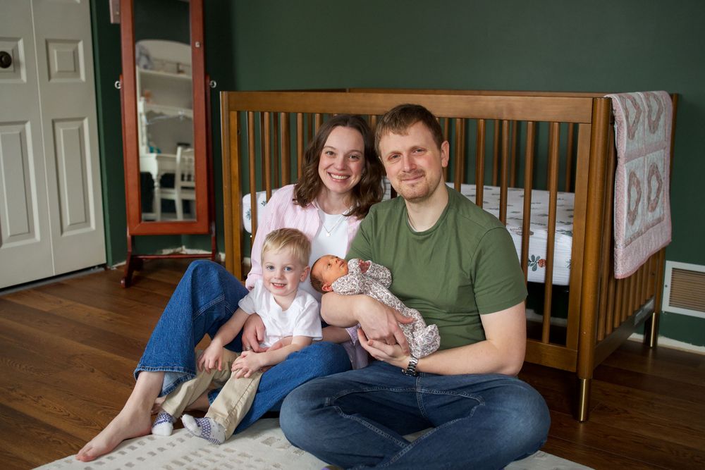 Family of four sitting on floor by wooden crib in green nursery, parents smiling with toddler and newborn baby.