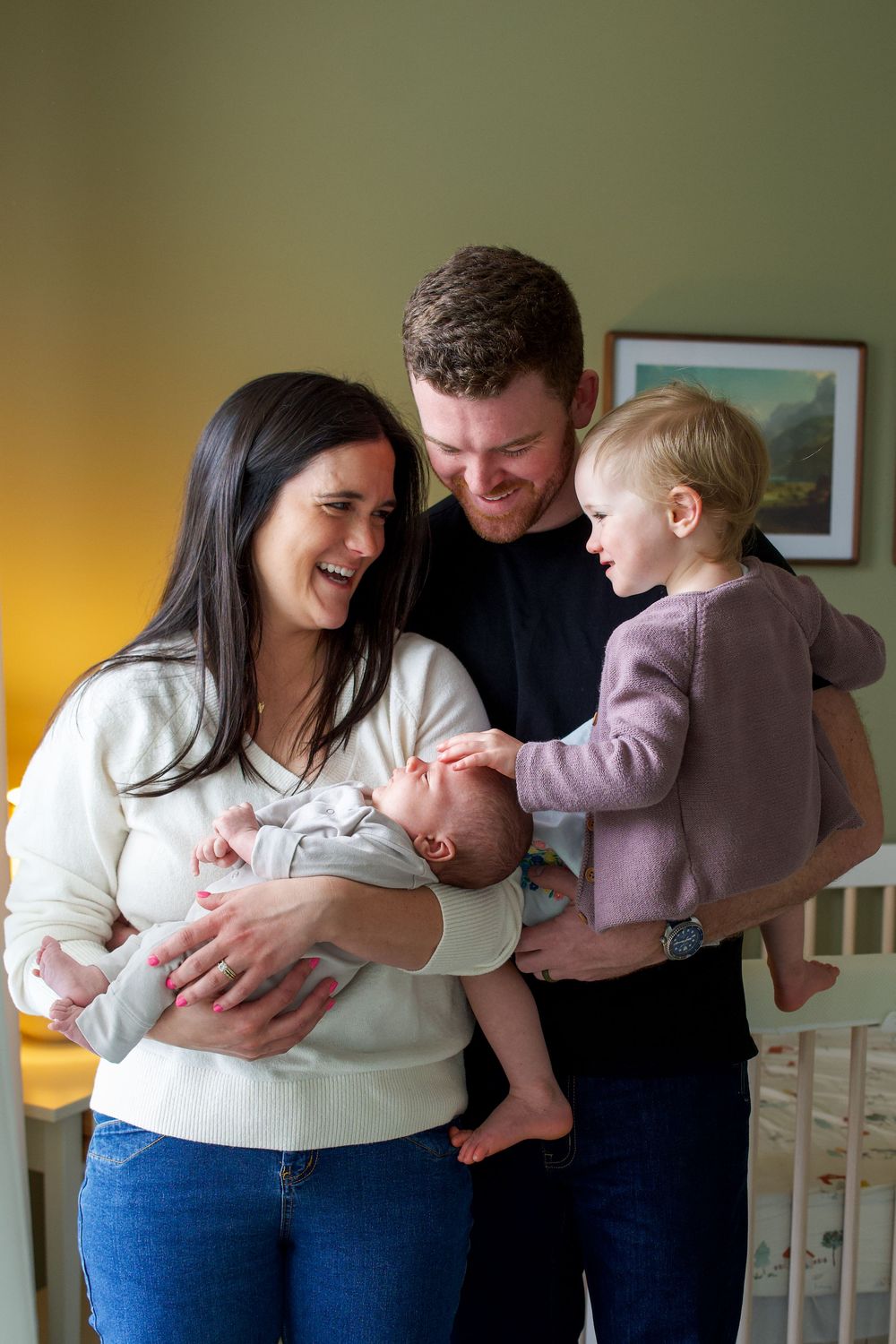 Happy family of four with parents holding newborn baby and toddler daughter in a cozy nursery room.