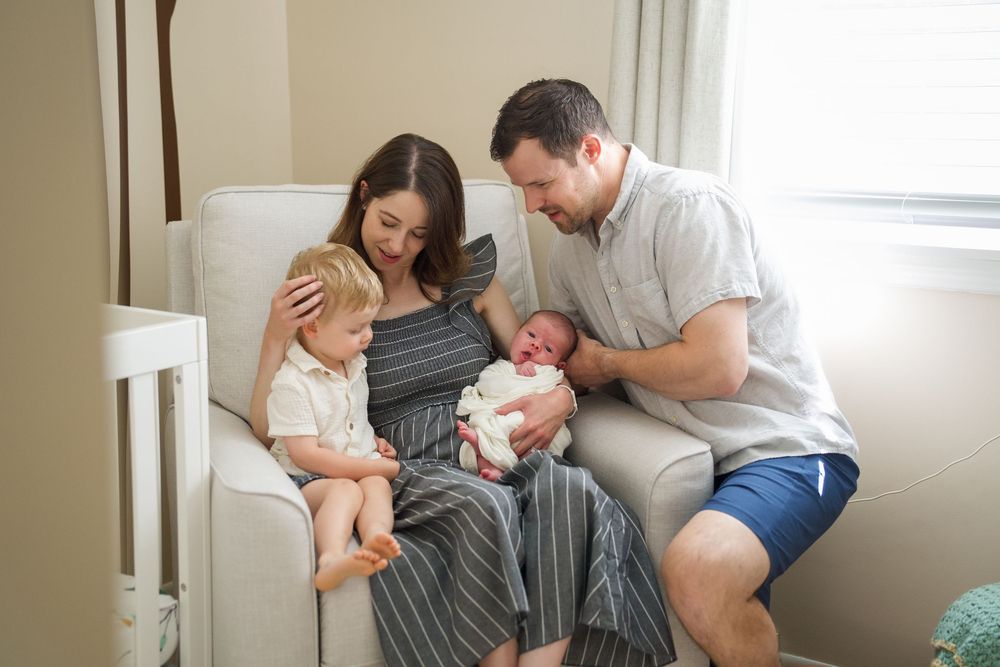 Family of four with newborn baby sitting in nursery chair, parents smiling at infant while toddler looks on.