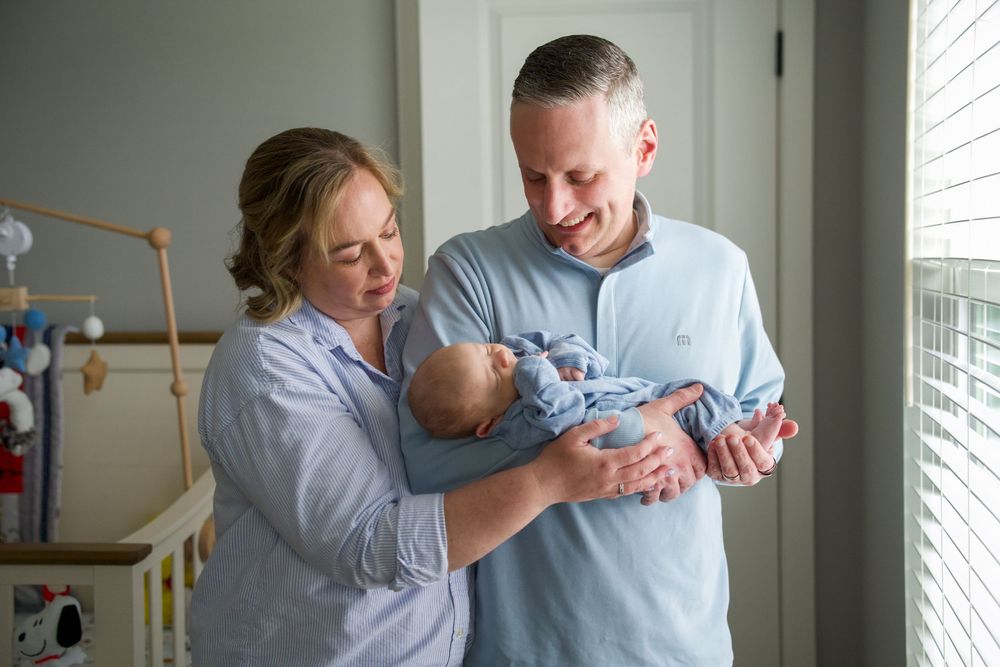 Happy parents smiling and holding their newborn baby in a nursery room near a window.
