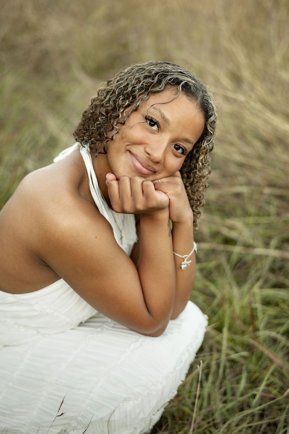 A person in a white dress poses thoughtfully in tall grass during an outdoor portrait session.