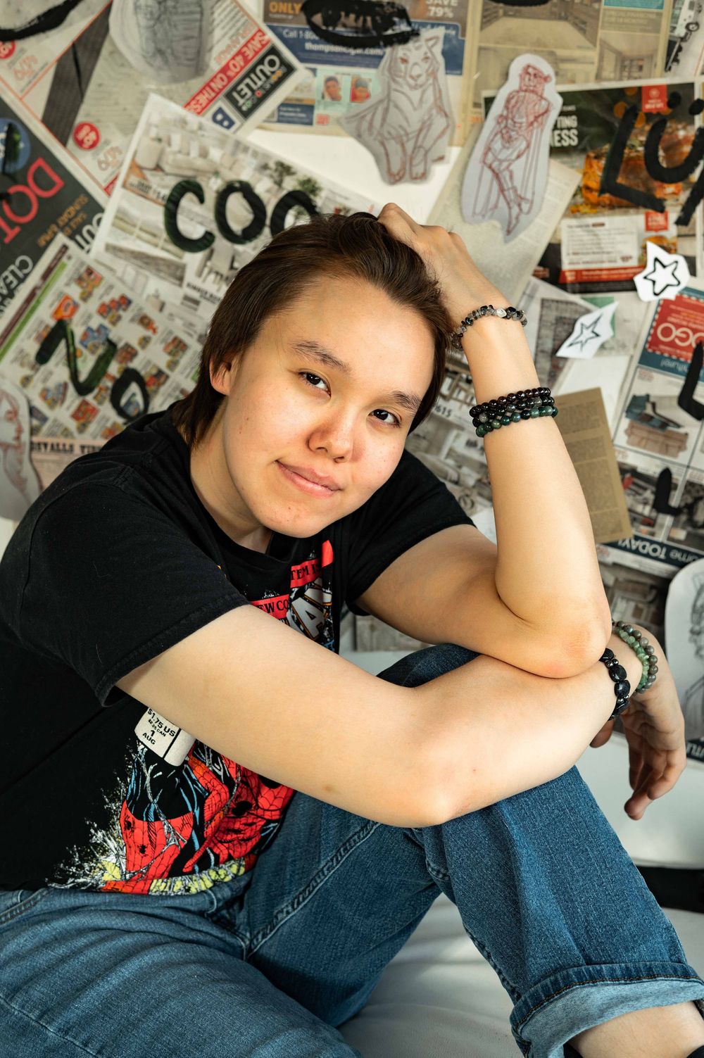 Young woman in black graphic tee and jeans sits relaxed against a wall covered in magazine clippings.