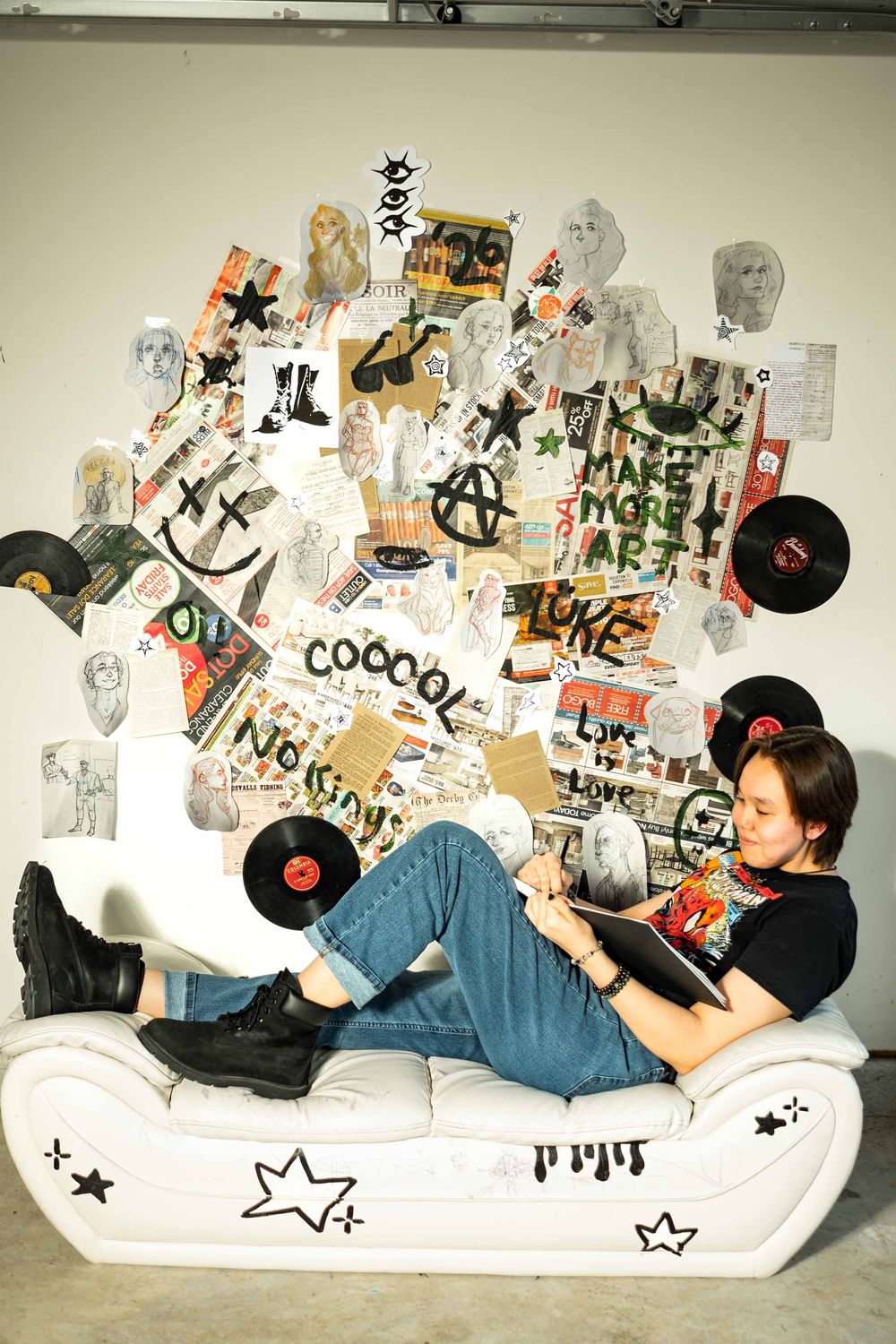 Person reclines on white sofa surrounded by vinyl records and eclectic collage wall art featuring punk-inspired graphics.