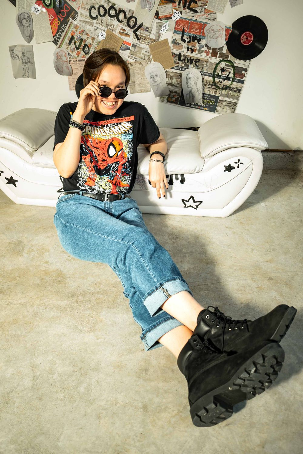 Young woman in Spider-Man tee, jeans, and black boots sits on floor wearing sunglasses in a decorated room.
