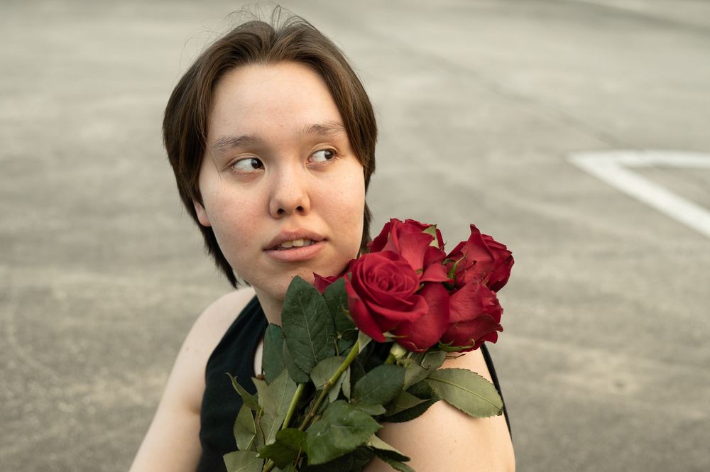 A person with dark hair holds vibrant red roses while looking to the side on a concrete background.