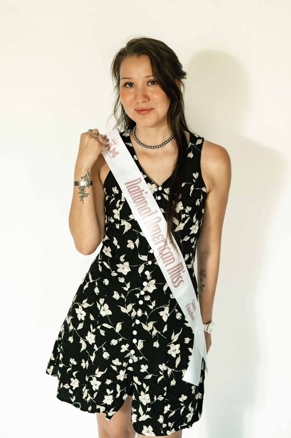 Young woman in black floral dress wearing a pageant sash, posing against white background.