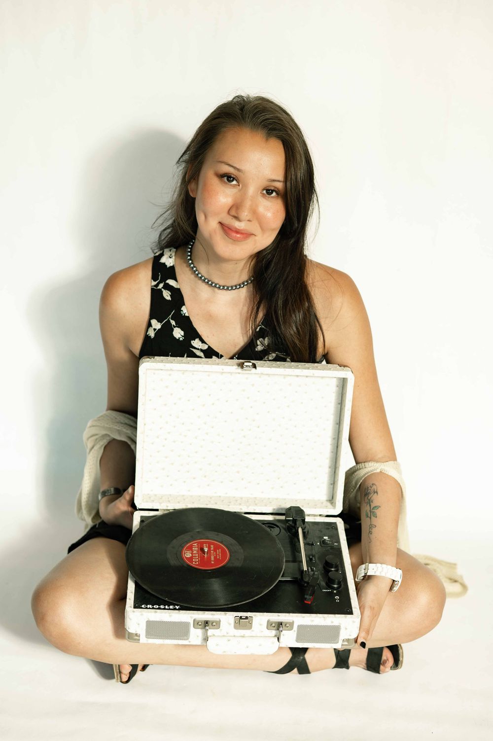 A smiling woman sits cross-legged holding a vintage portable record player with a vinyl record on a white background.