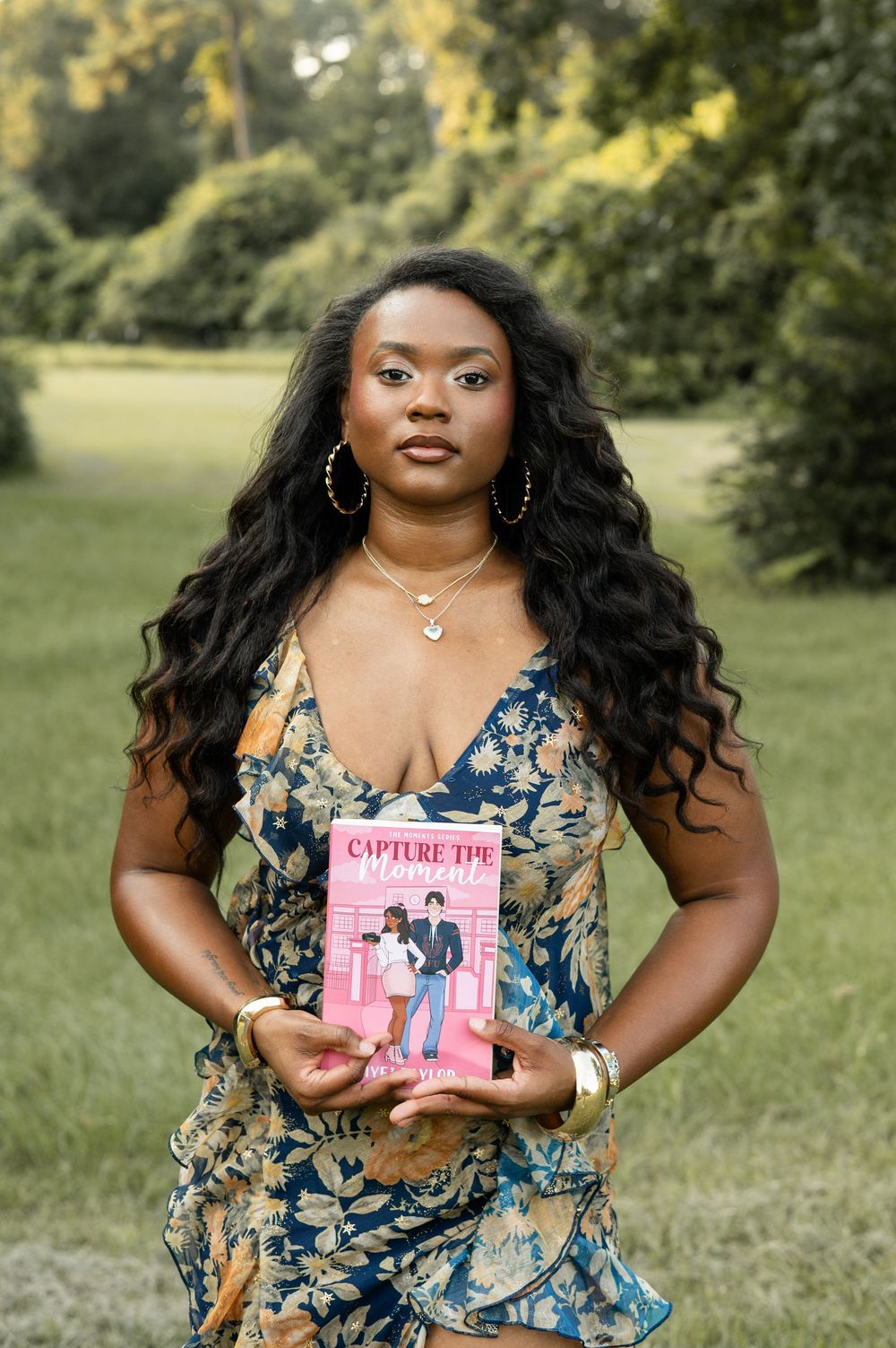 A person wearing a floral dress holds up a pink book titled 'Capture Life' while standing in a sunny outdoor setting.