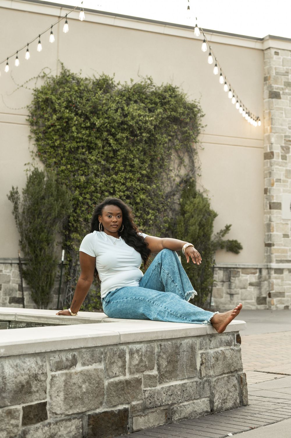 A person in blue denim lounges casually on a stone ledge outside a building with climbing vines.
