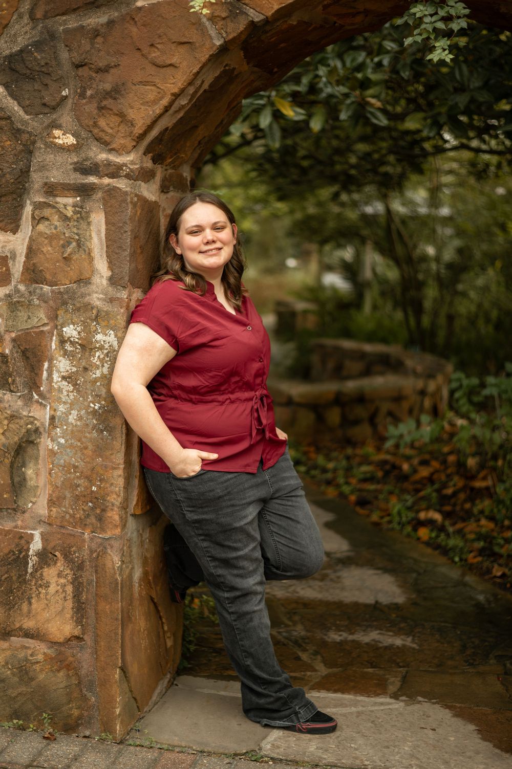 A person in a burgundy top and dark pants poses against a stone archway in an outdoor garden setting.