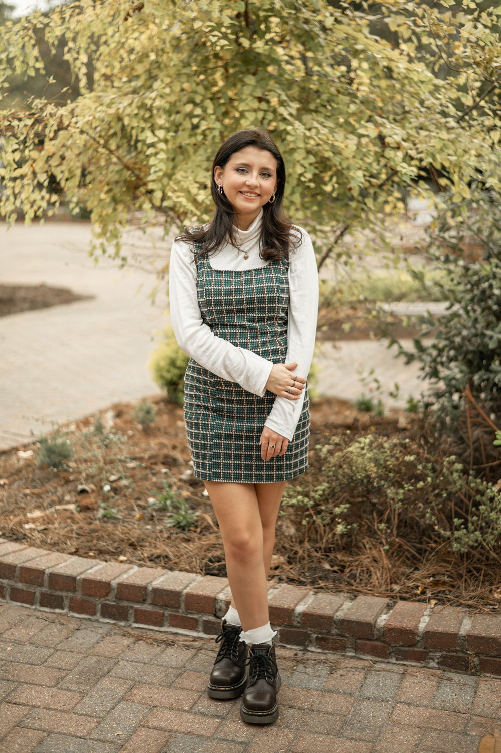A person wearing a plaid dress and white sweater poses outdoors near autumn foliage and a brick pathway.