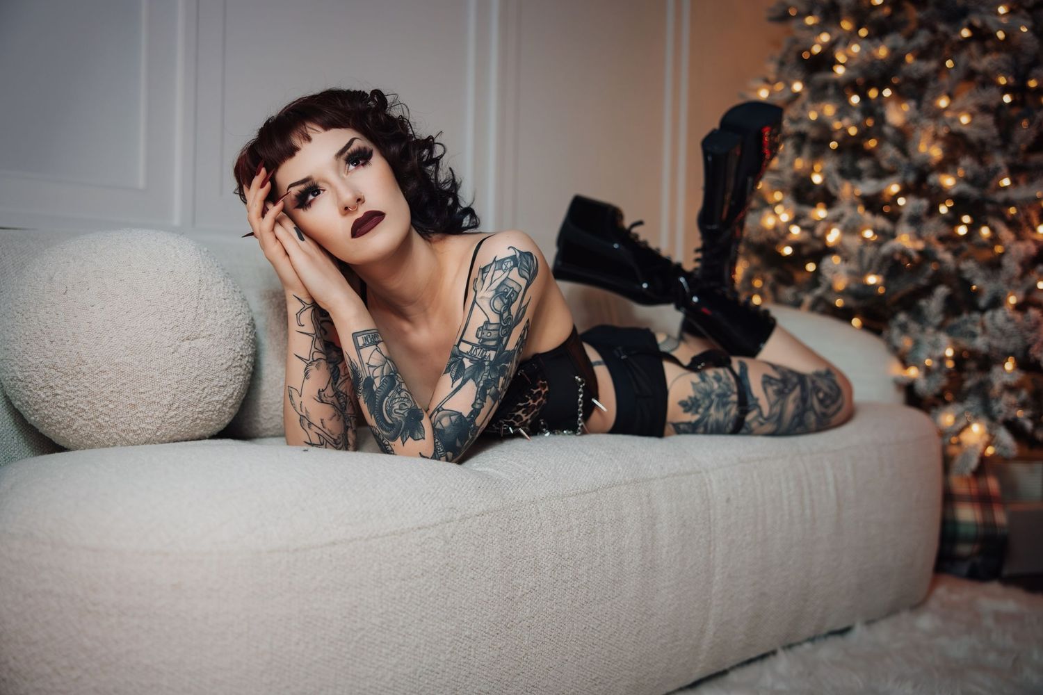 Person lounges on white couch near decorated Christmas tree with twinkling lights in cozy holiday setting.