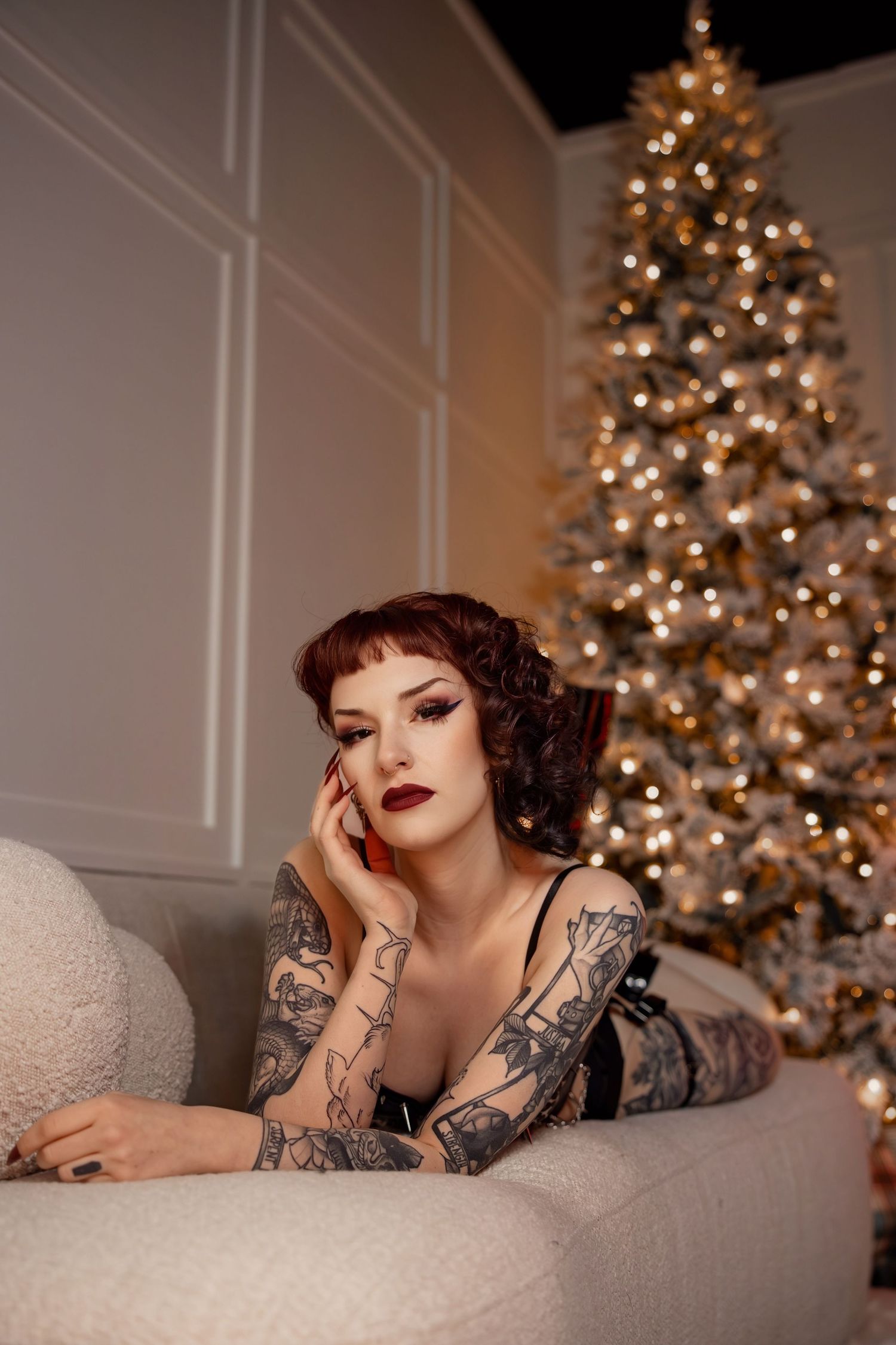 Woman in glamorous holiday photo shoot lounges on white couch with illuminated Christmas trees in background.