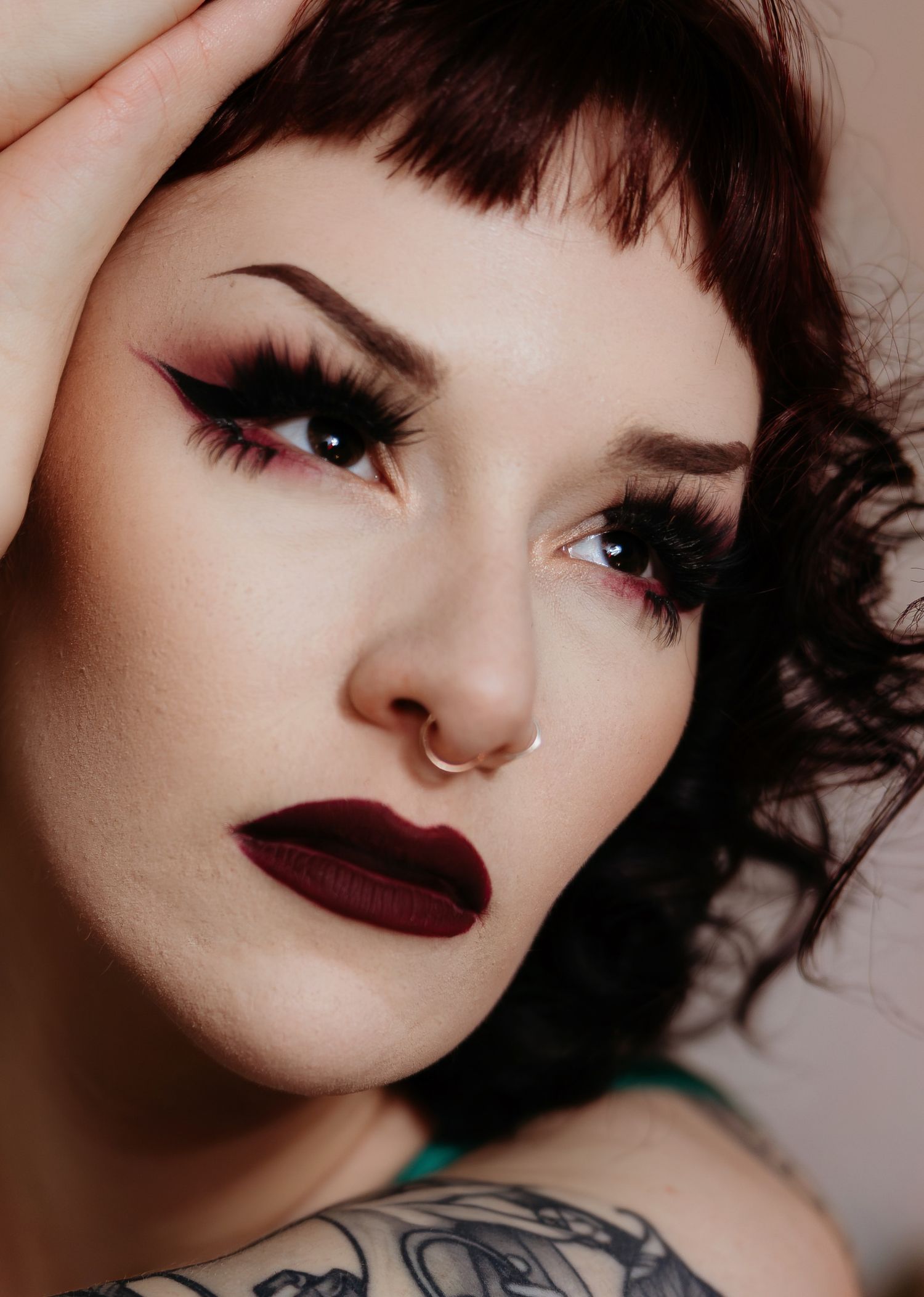 Close-up beauty portrait featuring dark plum lipstick, winged eyeliner, nose ring and short dark hair.