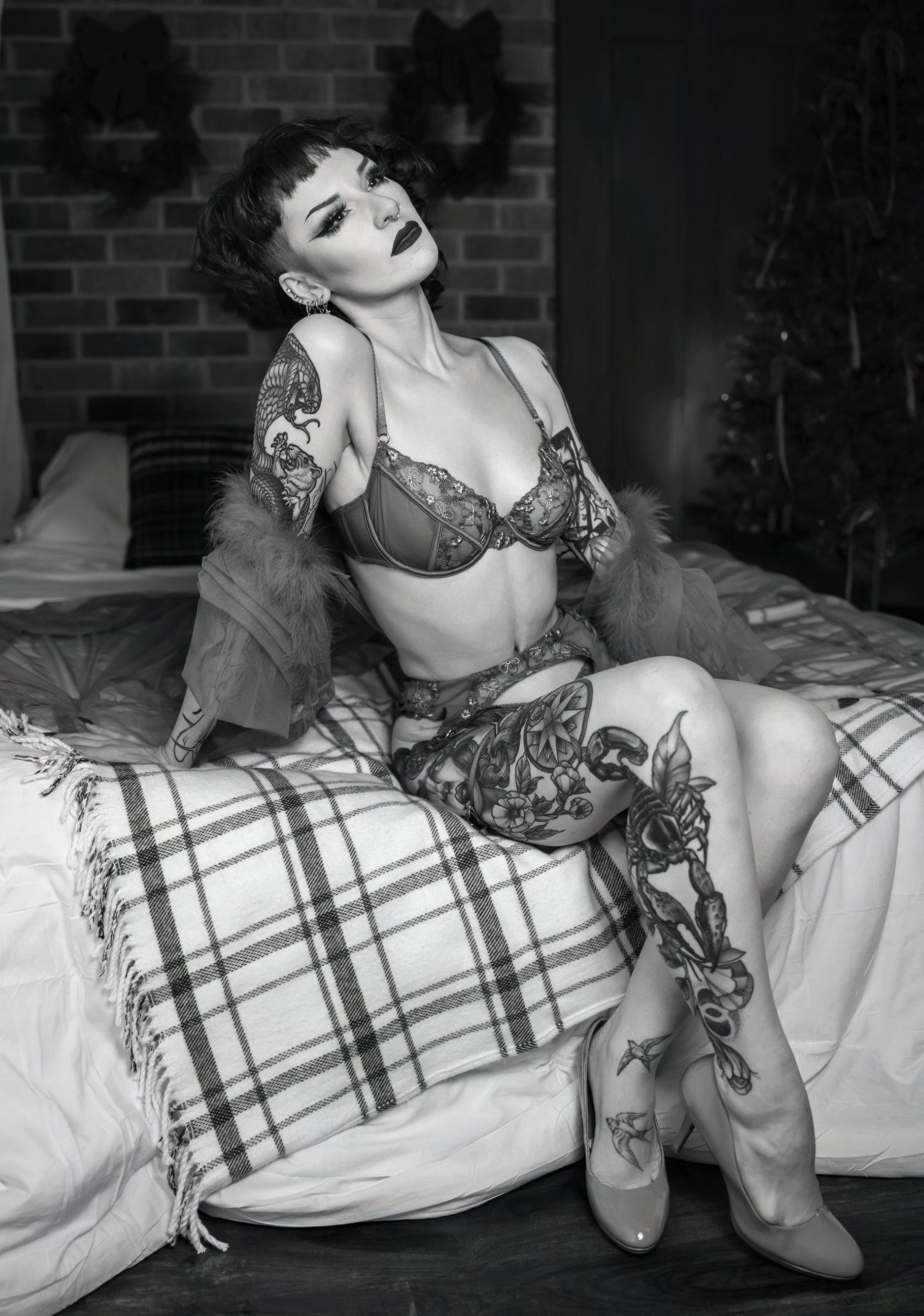 Monochrome boudoir photo showing a person in lingerie posing on plaid bedding with tattoos visible.