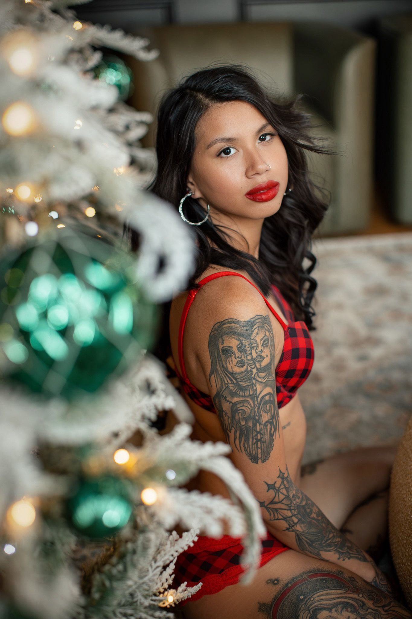 Woman in red dress with tattoos poses by Christmas tree with teal ornaments and white flocking decorations.
