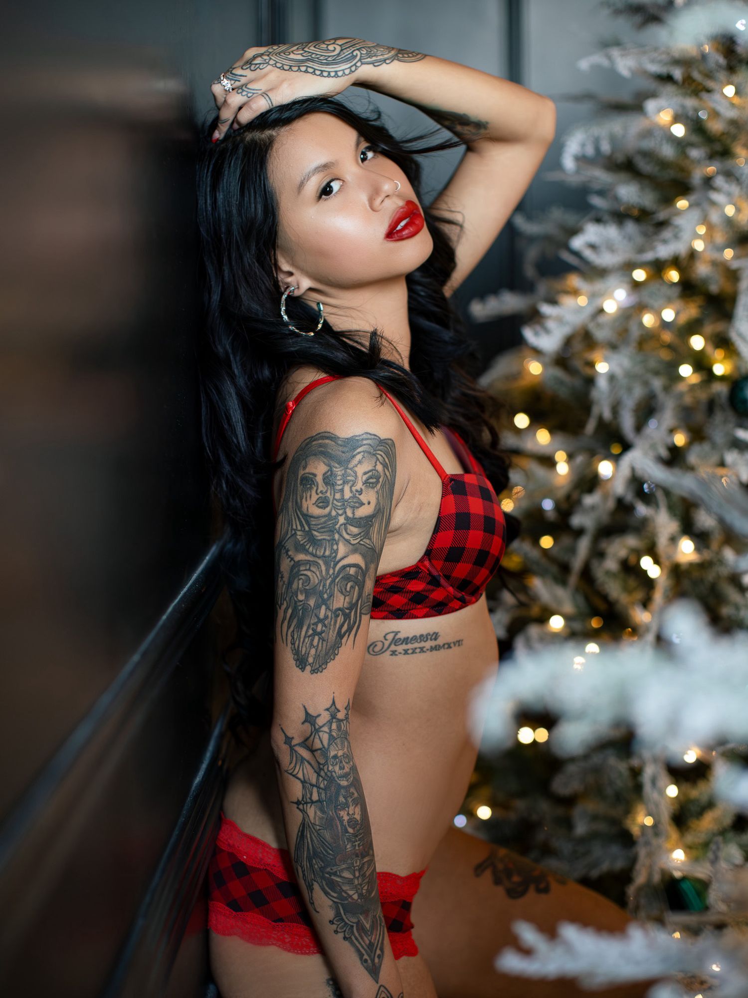 Festive holiday boudoir photoshoot featuring red plaid lingerie and arm tattoos next to a decorated Christmas tree.