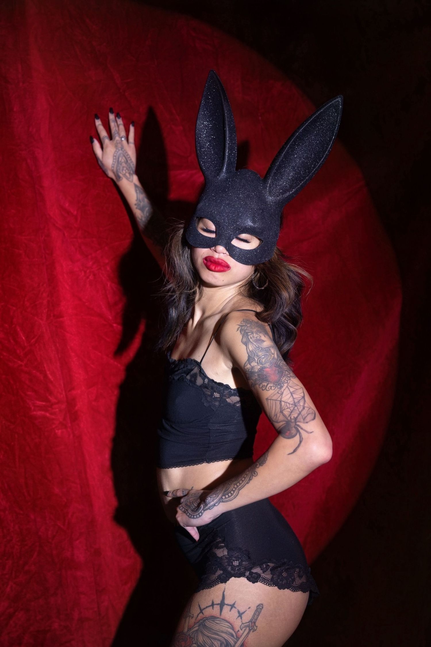 A figure in a dark bunny mask and tattoos poses dramatically against a deep red backdrop.
