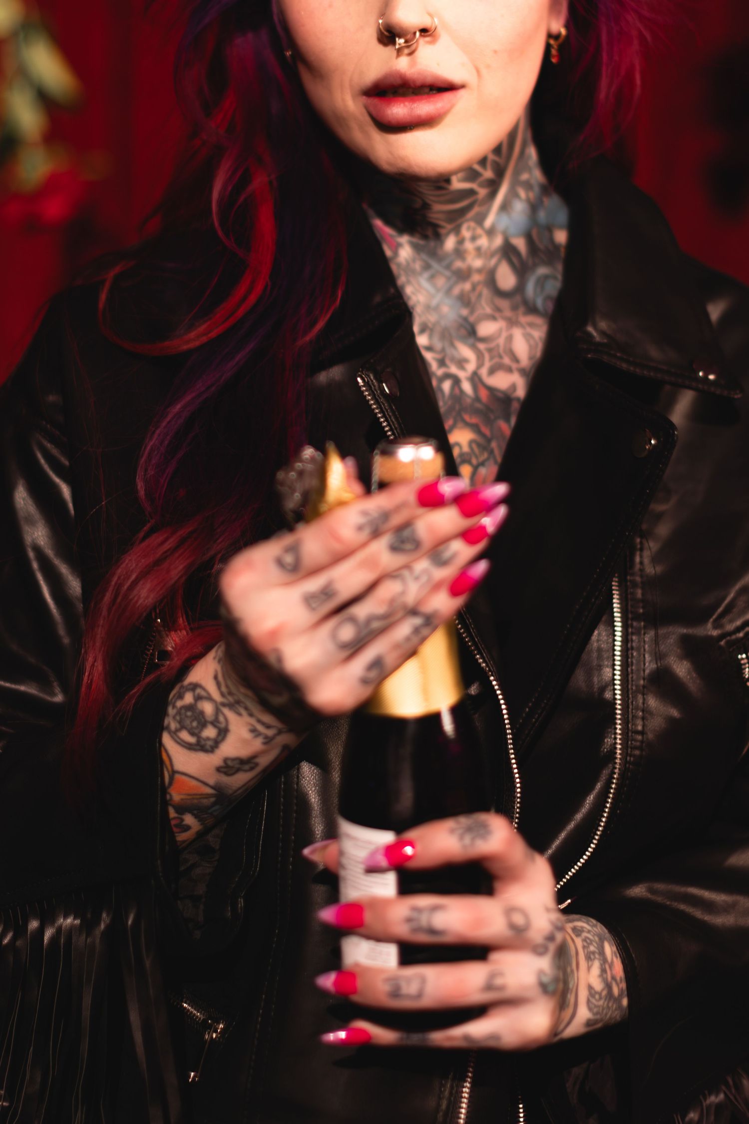 Tattooed hands with bright pink nails hold a beverage against deep red background.