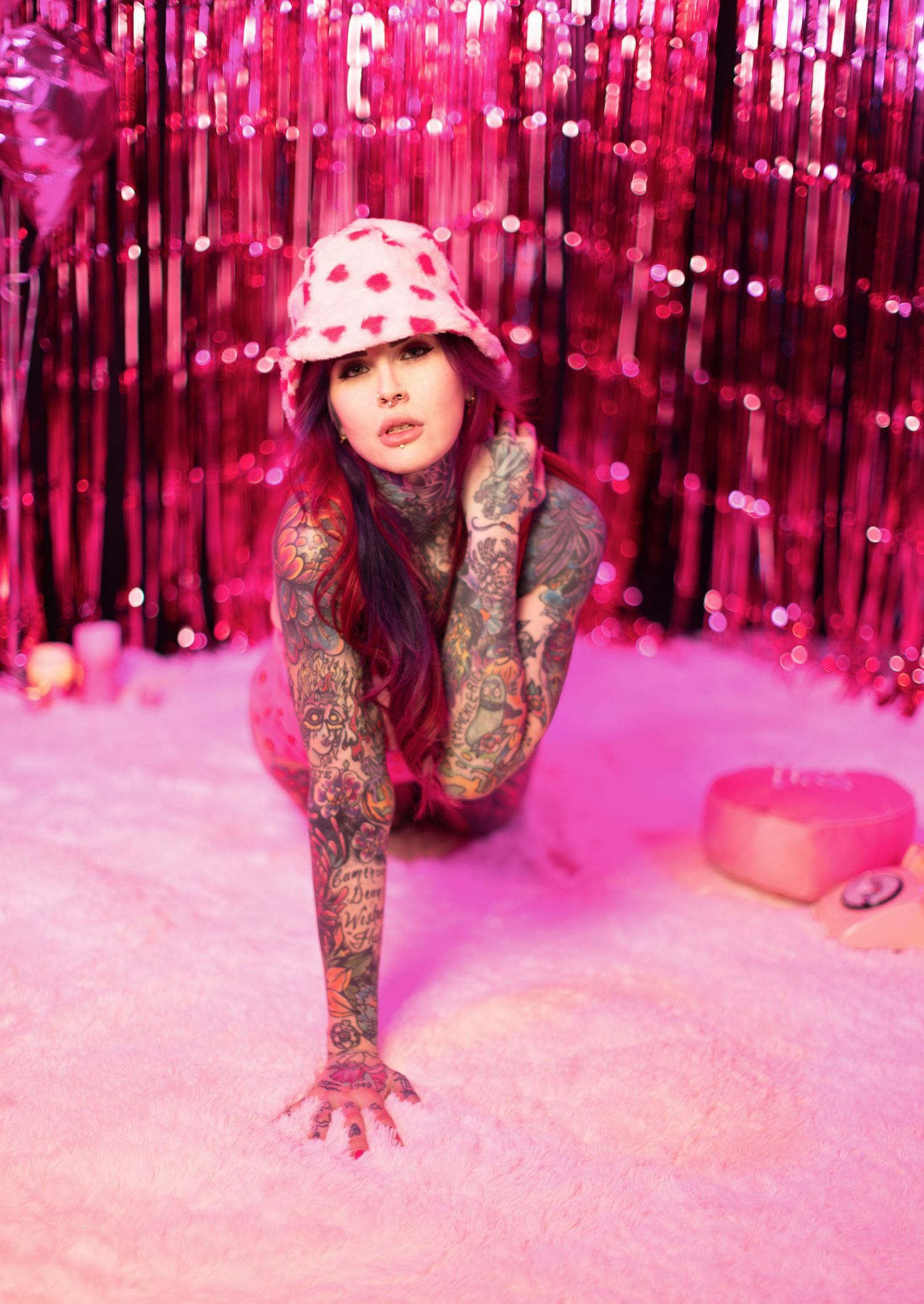 Model in alternative style poses against pink sparkly backdrop showcasing tattooed arms.