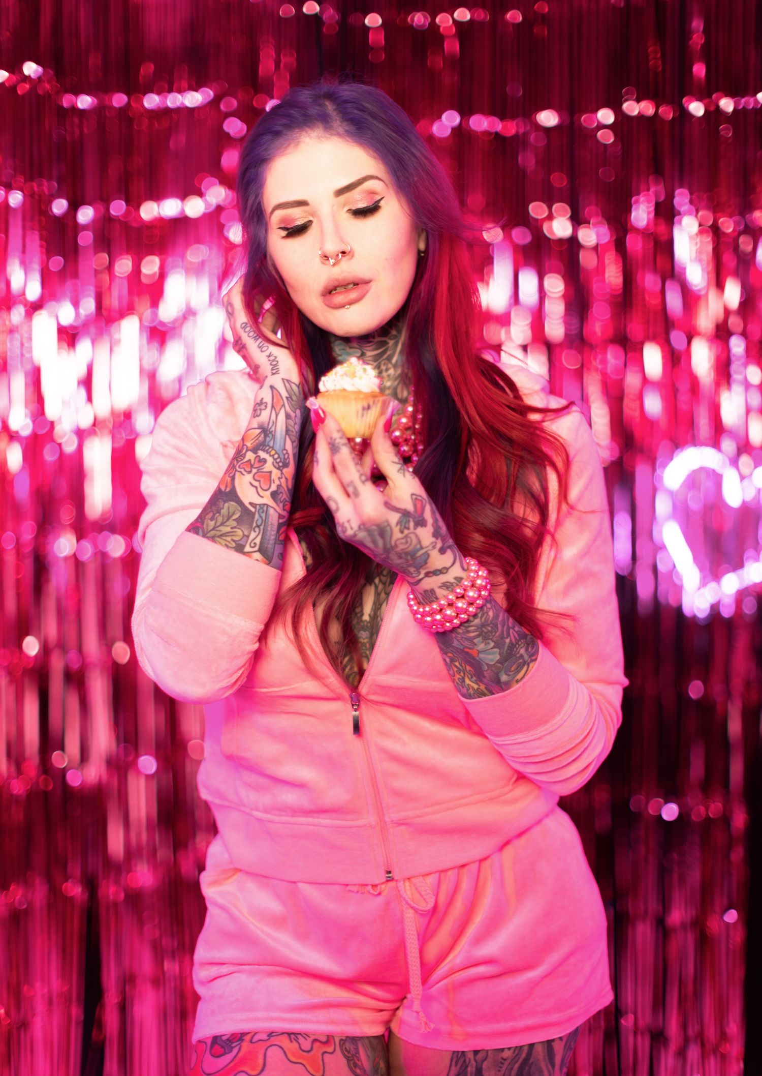 Person in pink sweater posing against shimmering pink backdrop with string lights creating a festive atmosphere.