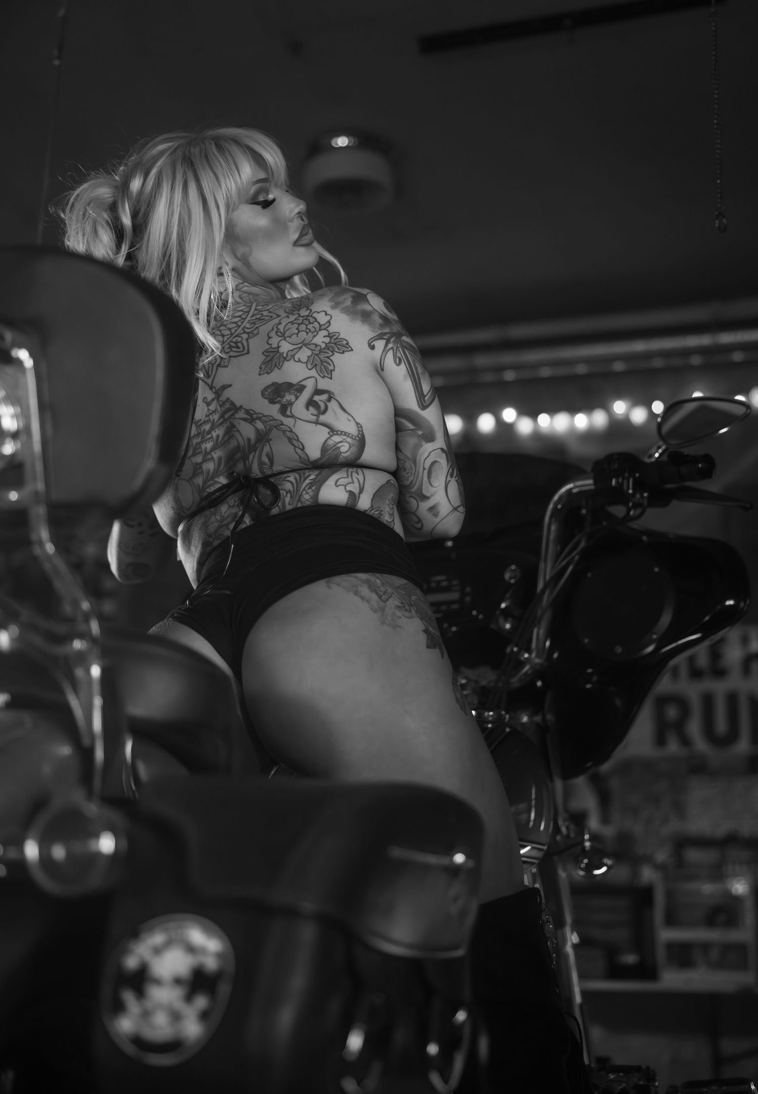 Black and white photo of a model posing on a motorcycle in a moody bar setting.