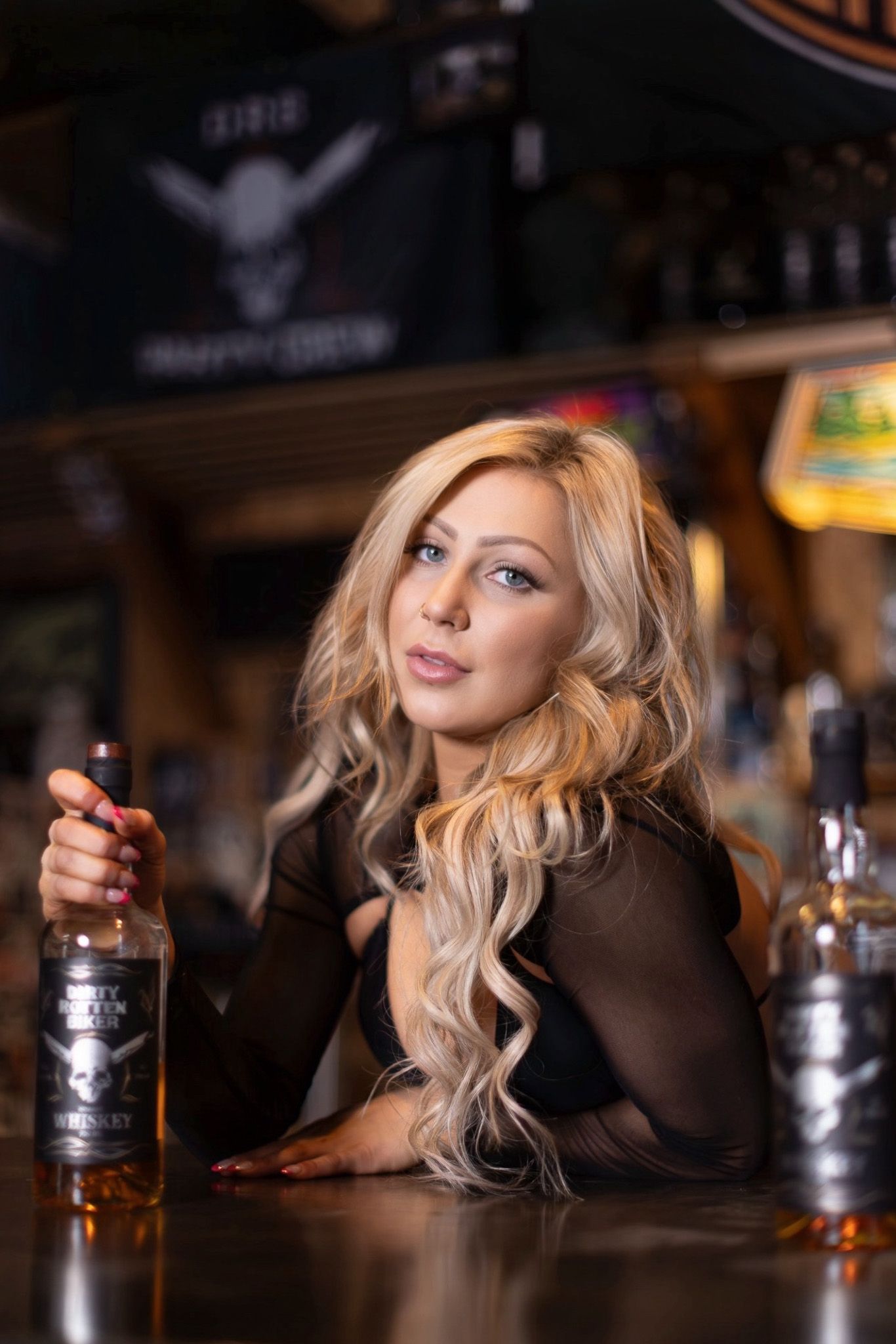 Blonde woman in black attire sits at bar counter with bottles of spirits in dimly lit establishment.