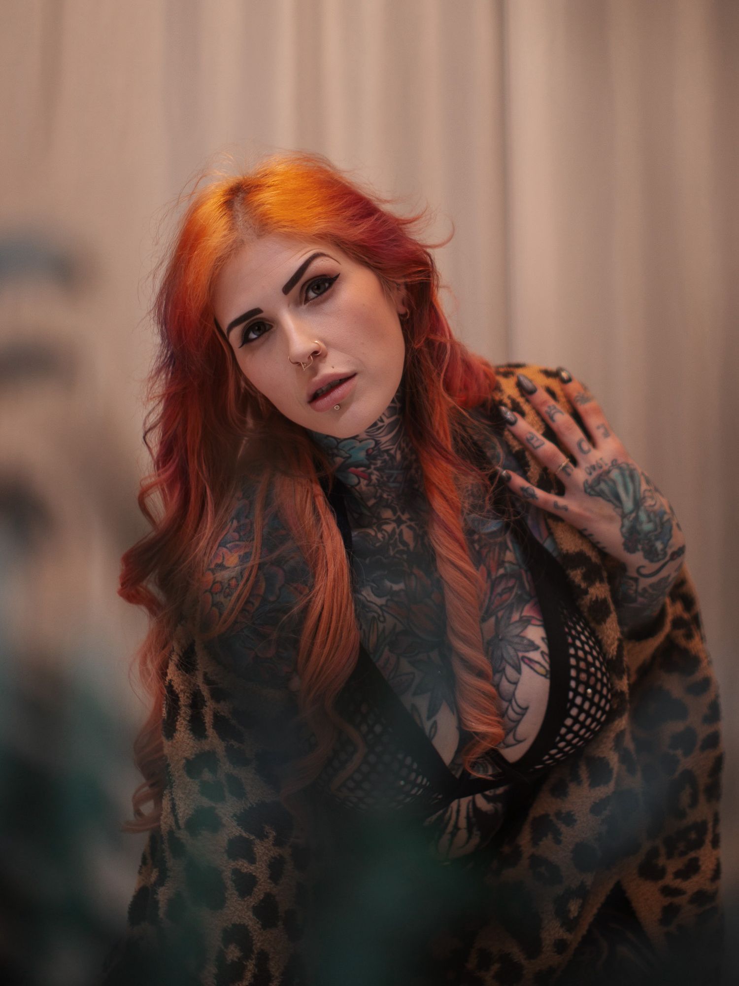 Woman with vibrant orange hair wearing a leopard print jacket and tattoos poses in moody lighting against a soft background.
