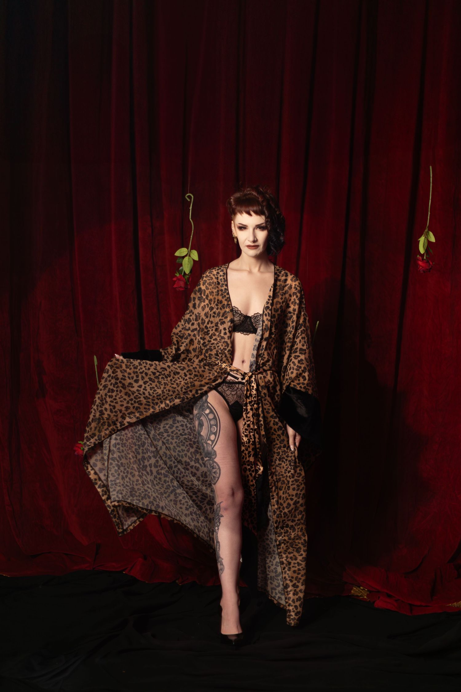 Figure in leopard print robe poses dramatically against red velvet curtain backdrop in vintage style photoshoot.