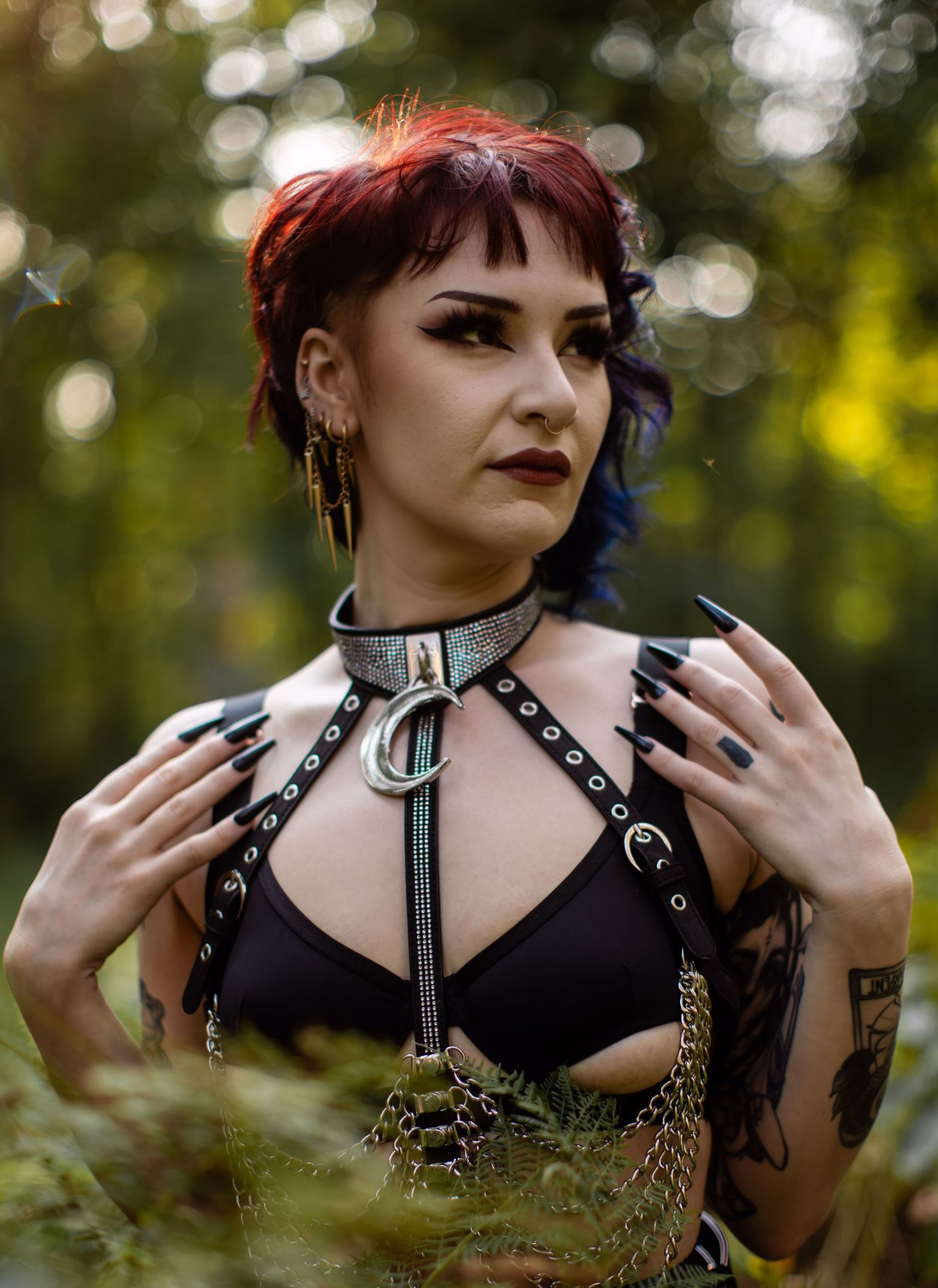Gothic model with leather harness and choker poses among ferns in natural lighting.
