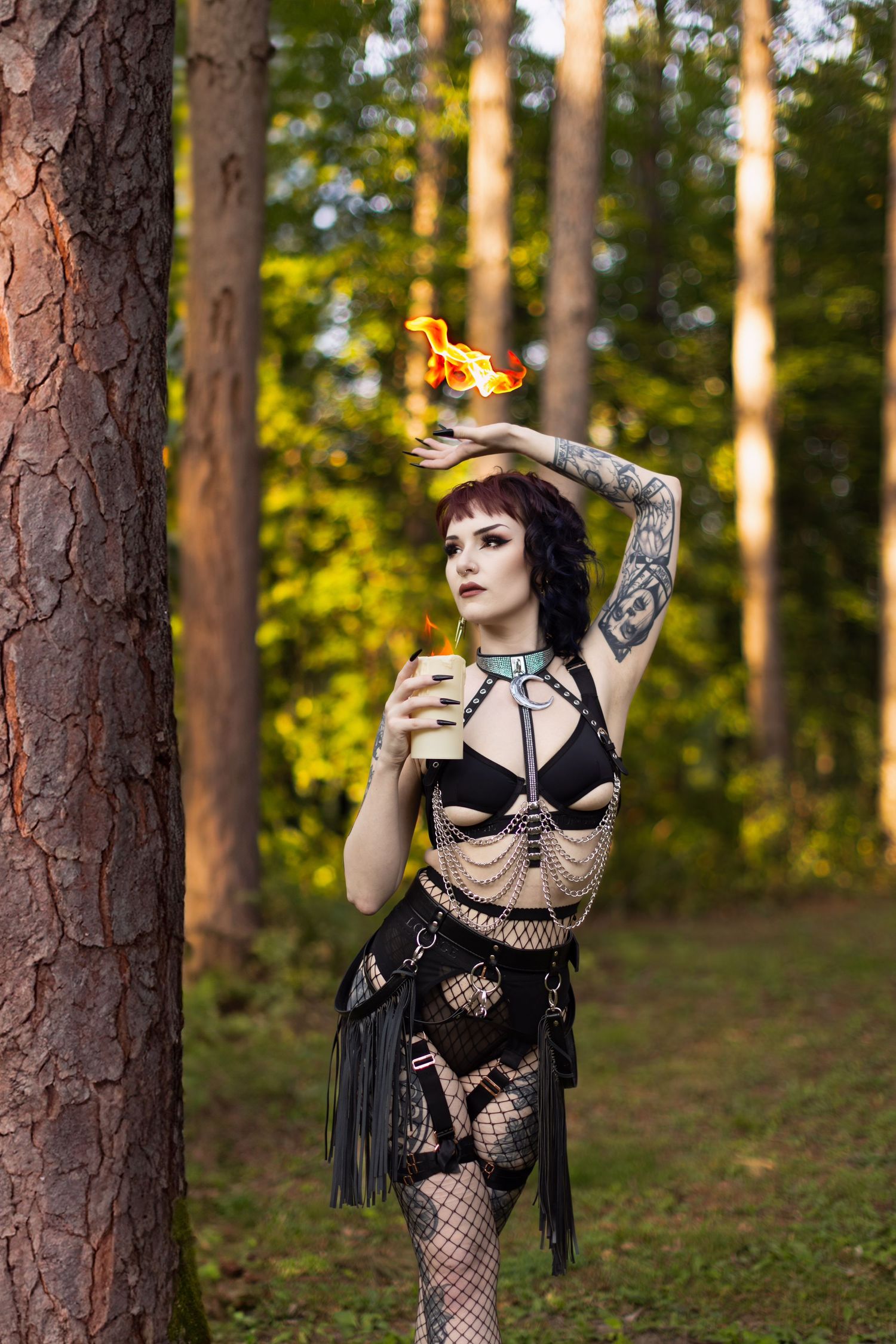 Gothic model in black outfit performs fire dance in forest setting.