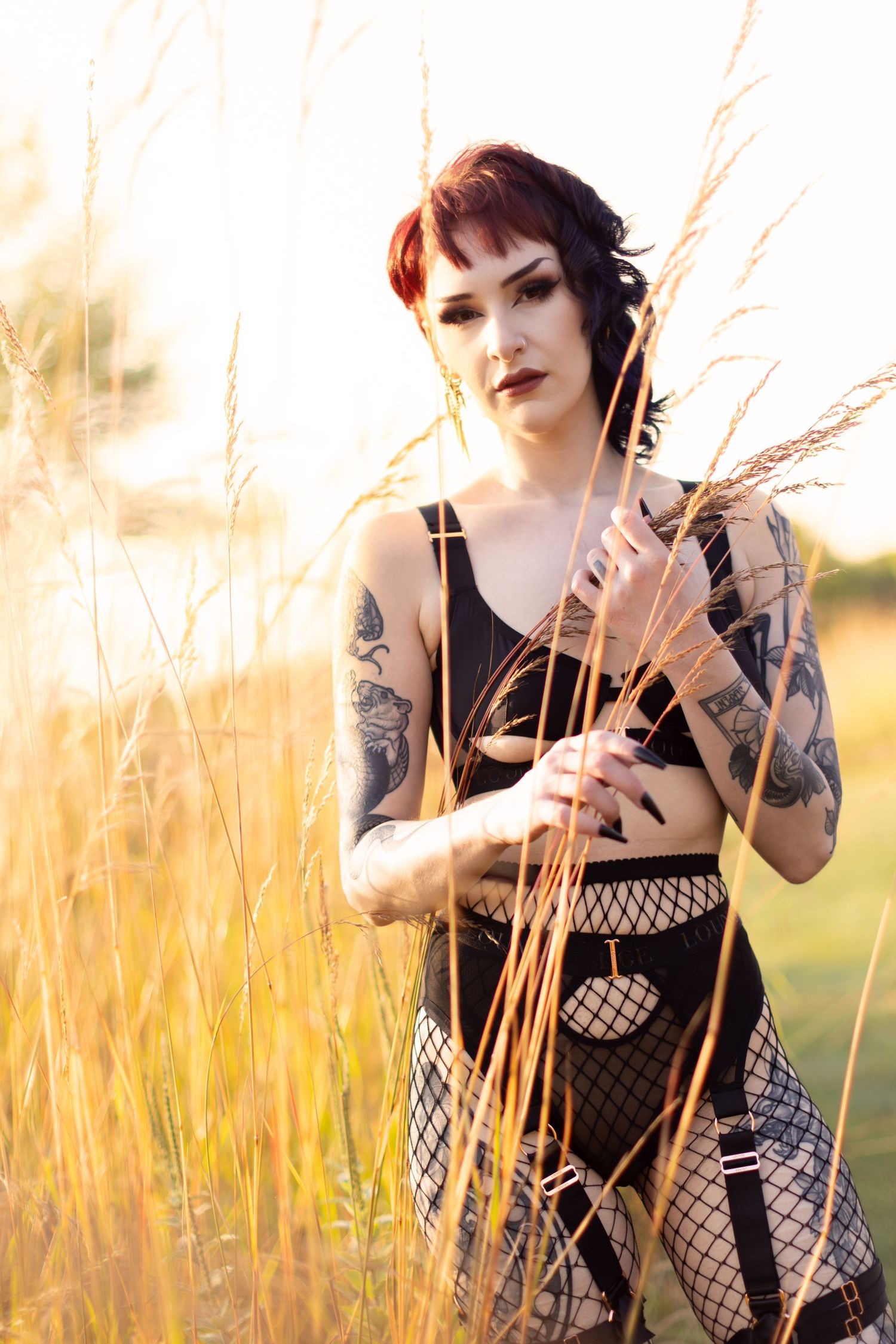 Alternative style model with tattoos poses in black outfit among golden wheat field.