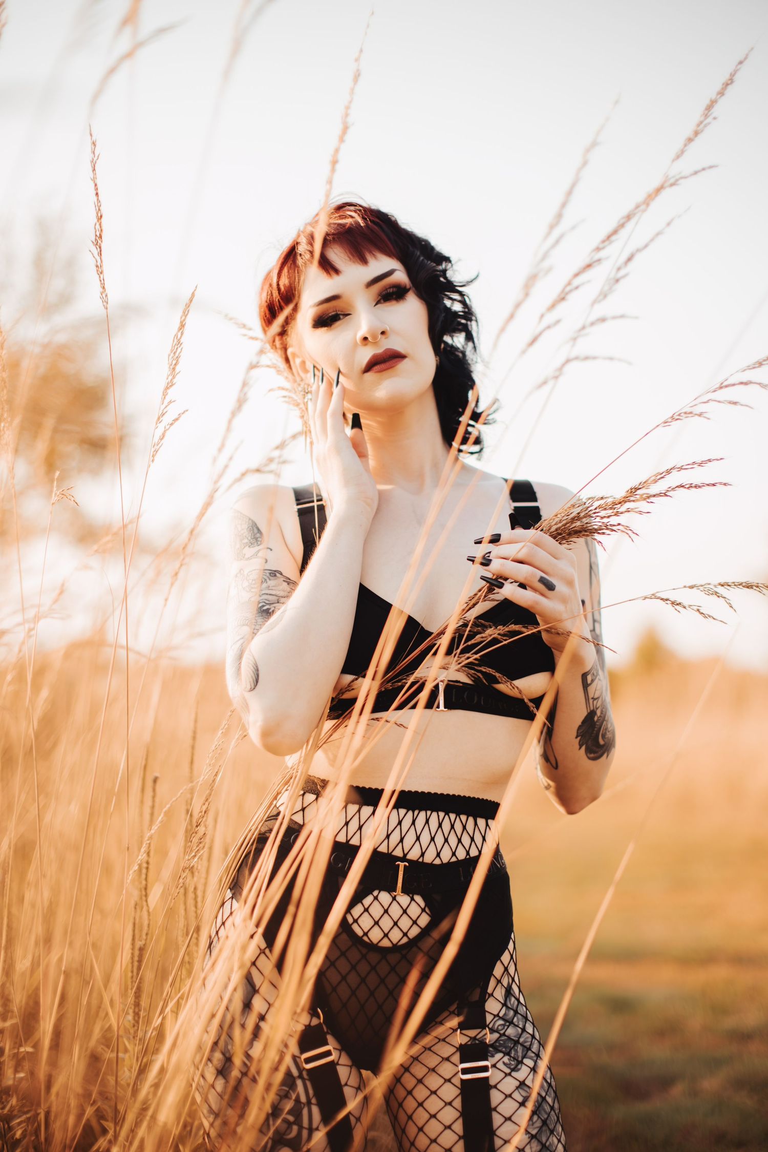 Model in black alternative outfit poses with dried grass during golden hour sunset.