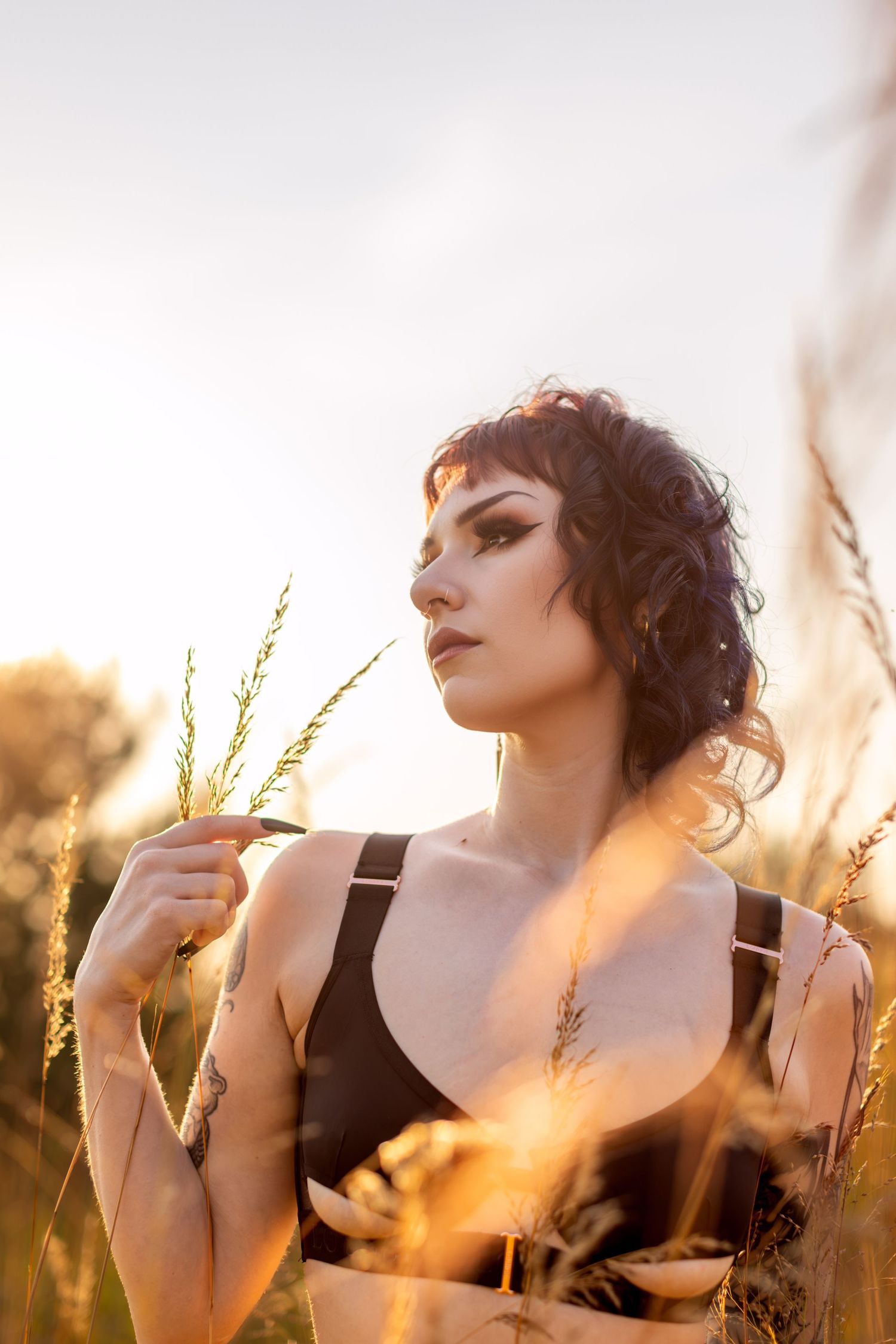 Dark-haired model poses in wheat field at golden hour wearing black crop top and holding wheat stalks.