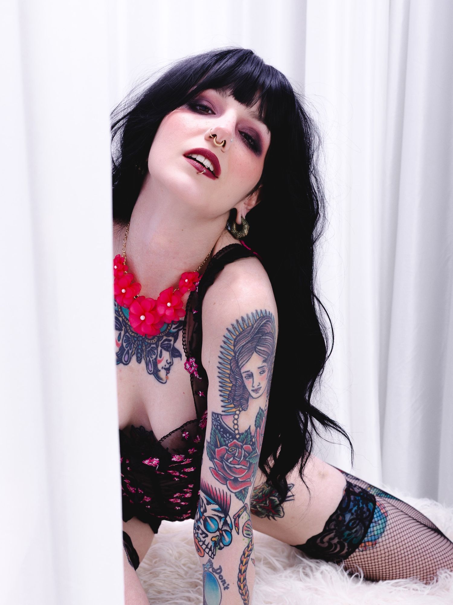 A model in black lingerie and red necklace poses against a white backdrop, showing off detailed arm tattoos.