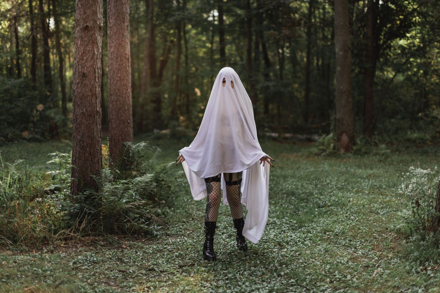 Ghostly figure in white sheet and boots walking through green forest path.