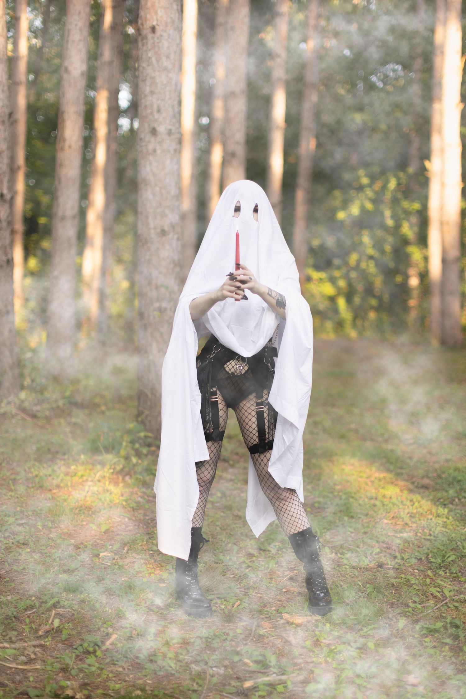 Ethereal ghost figure in fishnet stockings poses with red candle in misty forest.