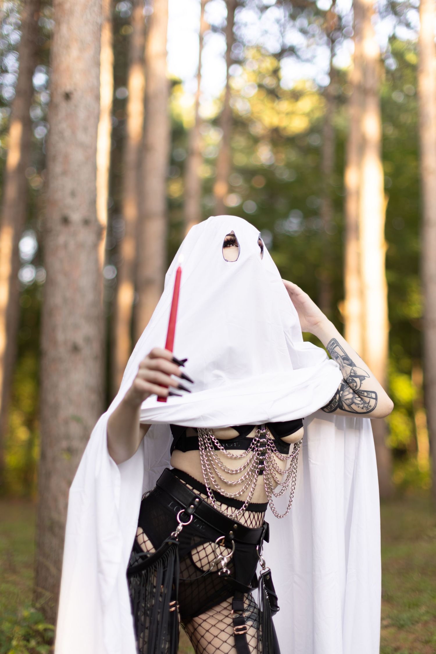 Ghost figure wearing white sheet and gothic attire holds red candle in wooded setting.