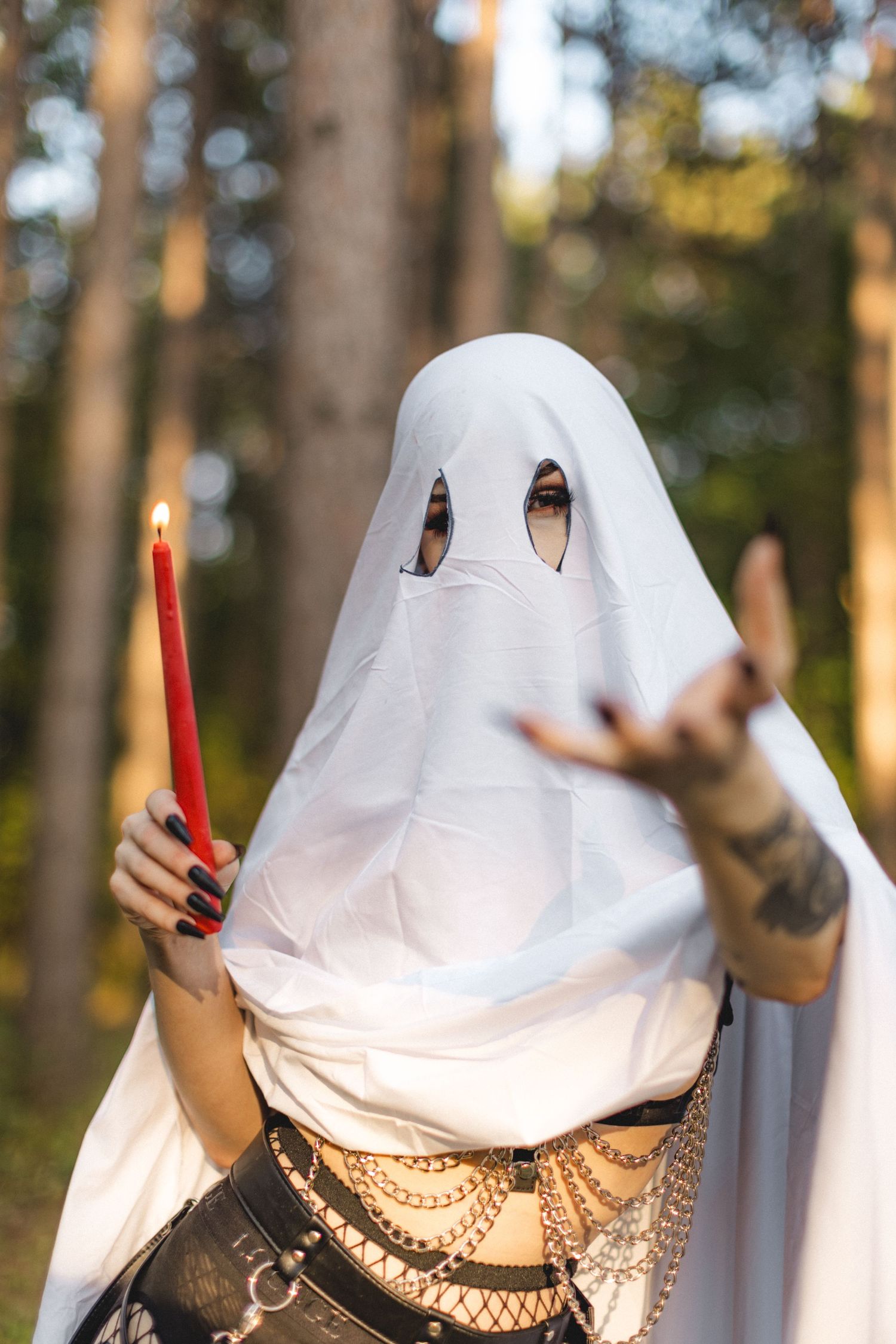 Spooky ghost costume with chain belt and red candle posing in sunlit forest.