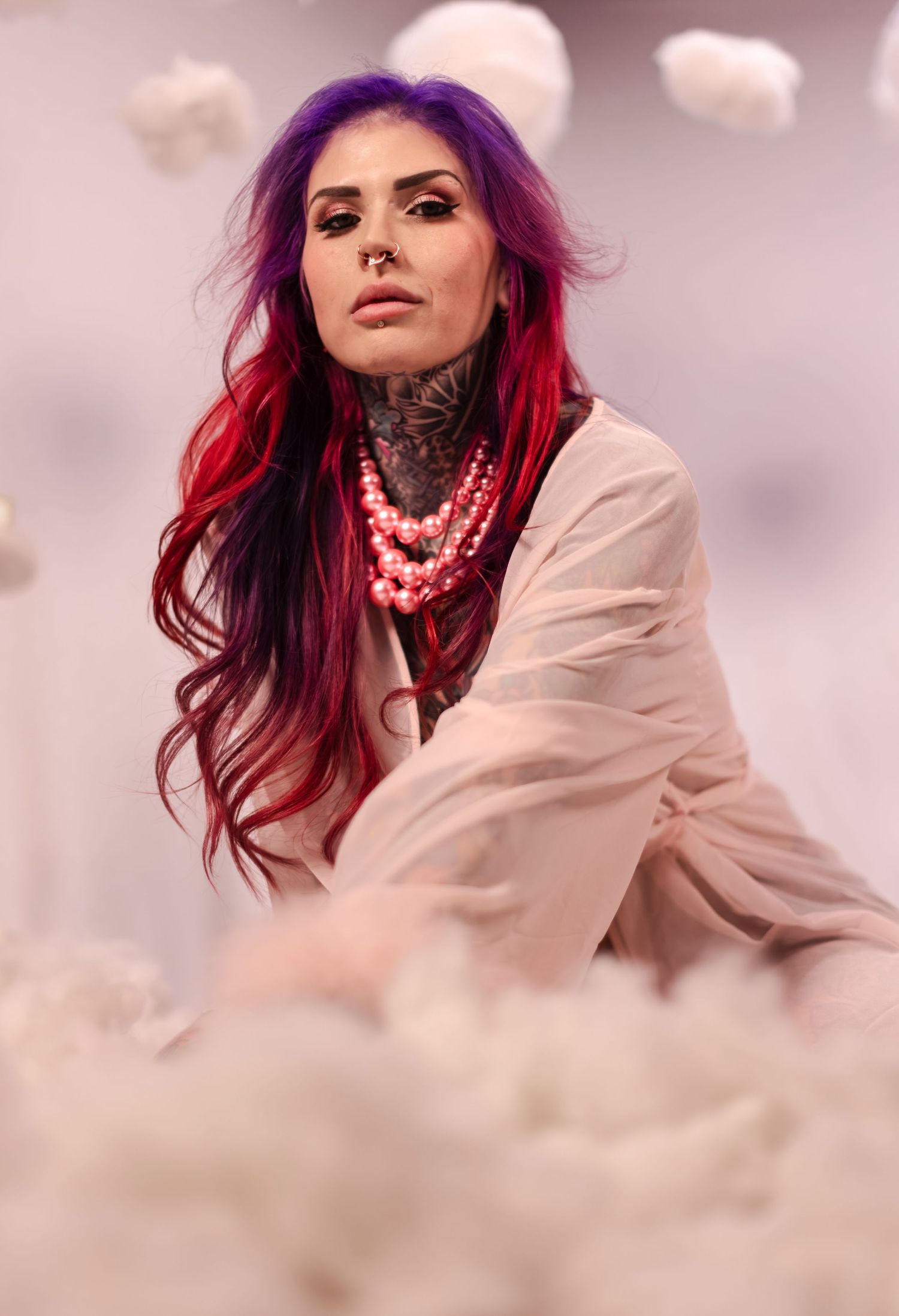 A model with vibrant red hair and white clothing poses ethereally against a dreamy pink-tinted cloudy sky backdrop.