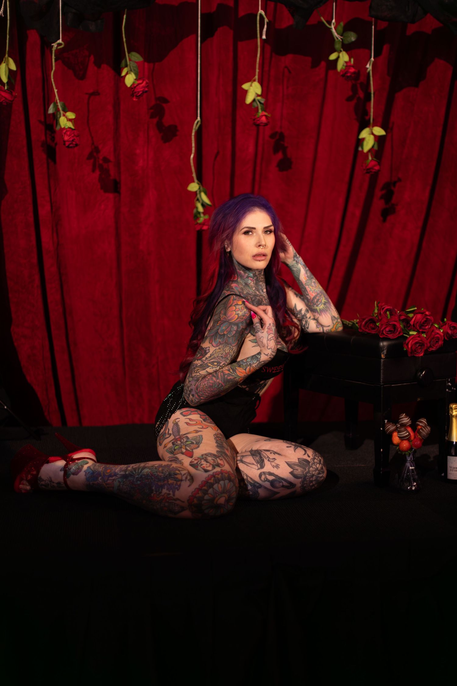 Tattooed model in black lace poses seductively against red curtain backdrop with hanging stars.