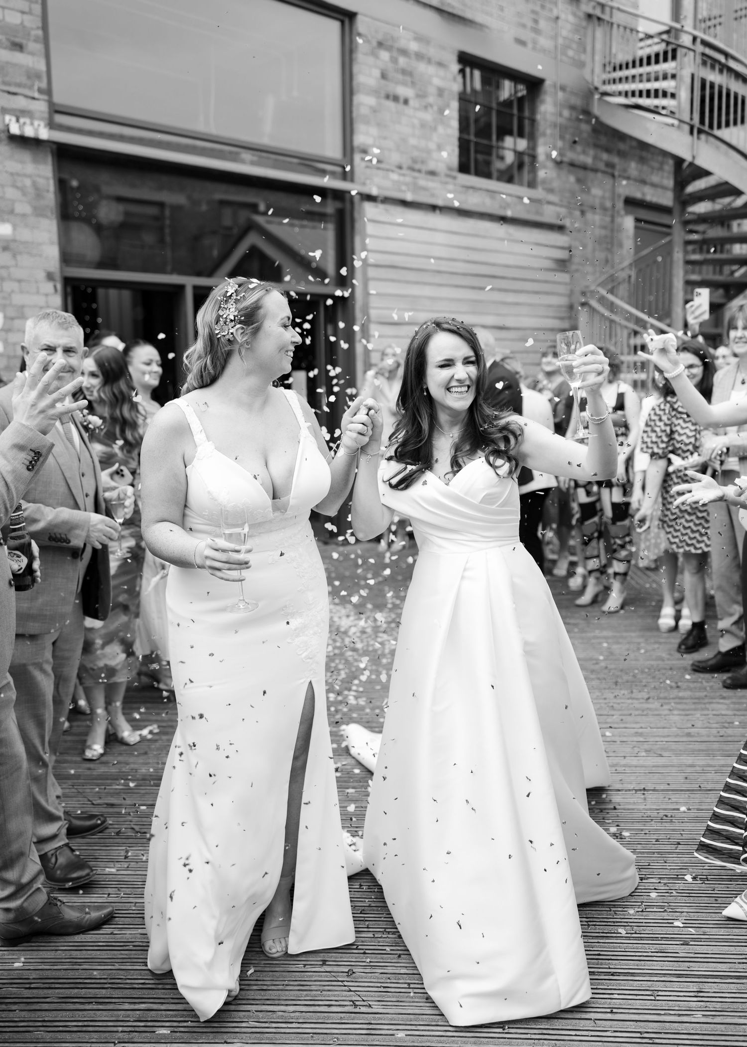 Two women in elegant white gowns celebrate outdoors while guests shower them with confetti in the UK.